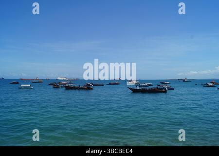 Les bateaux traditionnels en boutre en bois et les grands cargos flottent sur les eaux bleues scintillantes au large de Stone Town, Zanzibar. Banque D'Images