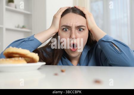 Jeune femme choquée regardant des cafards et des bretzels sur une table blanche à l'intérieur. Problème de ravageur Banque D'Images