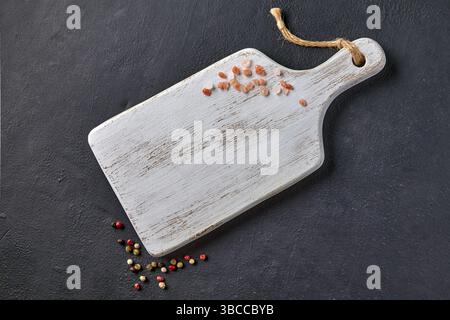 planche à découper blanche avec sel rose saupoudré et grains de poivre colorés sur la table en pierre noire Banque D'Images