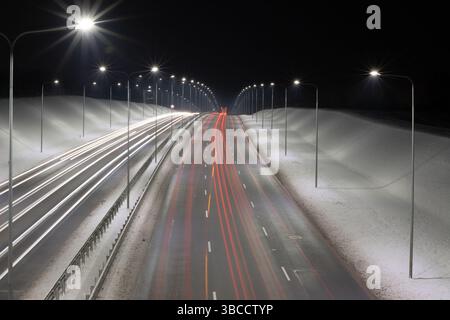 Une vue nocturne sereine d'une autoroute à plusieurs voies encadrée par des remblais enneigés et des lampadaires lumineux. Capturé avec une exposition longue Banque D'Images
