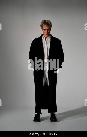 Portrait en pied d'un jeune homme élégant dans un manteau noir et un short blanc posant sur un fond gris Banque D'Images