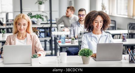 Travailler dans un bureau moderne, la technologie numérique et la distanciation sociale après la quarantaine. Femmes souriantes européennes et afro-américaines travaillant sur des ordinateurs portables, s'assoit Banque D'Images