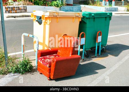 Fauteuil vintage abandonné placé près de bacs de recyclage colorés dans une rue de la ville. Décomposition urbaine, élimination des déchets et thèmes environnementaux Banque D'Images