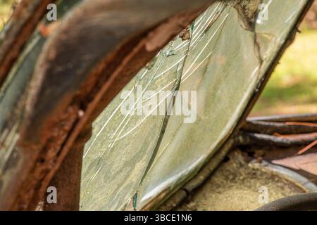Gros plan d'une fenêtre de voiture fissurée encadrée par du métal rouillé et un rembourrage délavé, soulignant l'âge, la décomposition et l'histoire automobile oubliée. Banque D'Images