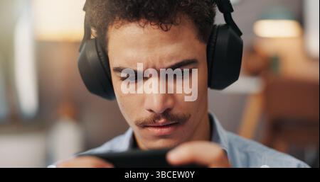 En ligne, jeux vidéo et homme avec écouteurs, smartphone et sérieux pour le sport virtuel dans l'appartement. Streaming, joueur et personne avec application mobile pour Banque D'Images