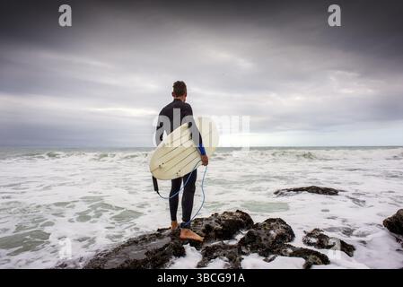 Un surfeur se tient sur les rochers avec sa planche de surf sous le bras alors qu'il regarde le surf juste avant de sauter dans l'eau Banque D'Images