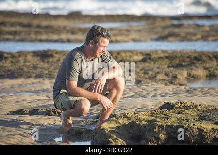 Un homme d'âge moyen est assis sur ses hanches sur les rochers près de l'océan tout en regardant vers le bas vers les rochers Banque D'Images