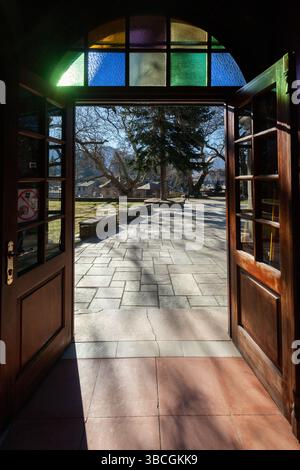 La porte en bois ouverte d'une église avec des vitraux révèle une cour en pierre ensoleillée et des arbres dans le village traditionnel de Metsovo, en Grèce. Banque D'Images