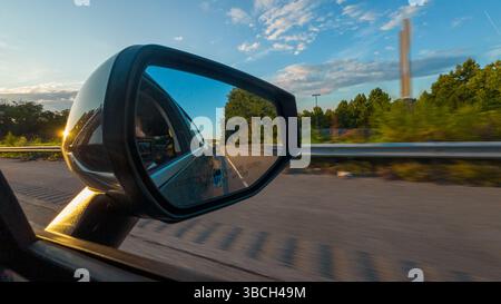 Une voiture roule sur une route avec une personne sur le siège du conducteur. Le rétroviseur de la voiture montre un reflet des arbres et du ciel en arrière-plan Banque D'Images