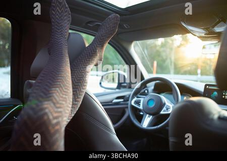 Jambes en collants à motifs reposant sur le siège d'auto, intérieur ensoleillé, volant visible. Banque D'Images