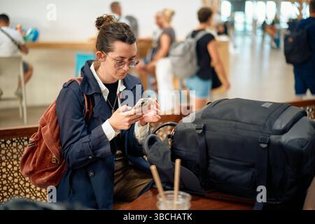 Une jeune femme avec un sac à dos vérifie son téléphone dans un terminal d'aéroport animé. Banque D'Images