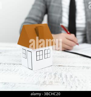 Femme d'affaires assise et écrivant dans un cahier. Maison de papier sur le bureau Banque D'Images