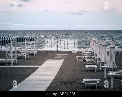 Une plage vide à Grado, en Italie, avec des rangées soignées de chaises longues et des parasols fermés flanquant une passerelle en bois qui mène directement à la mer sous une humeur changeante, Banque D'Images