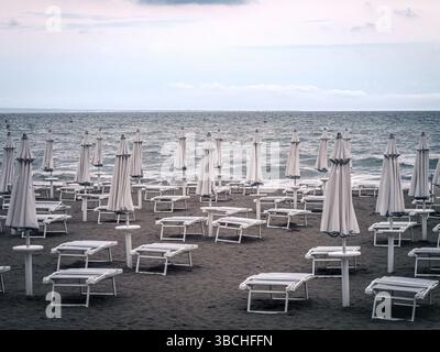 Une plage déserte à Grado, en Italie, avec des rangées de chaises longues blanches et des parasols fermés sous un ciel nuageux. L'atmosphère calme et hors saison contraste avec t Banque D'Images