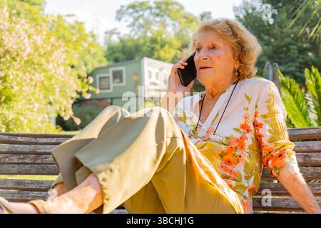 Femme âgée profitant d'une conversation sur son téléphone portable tout en se relaxant sur un banc en bois dans un jardin paisible, entouré de verdure et BA Banque D'Images