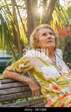 Femme senior profitant d'un moment de tranquillité dans un jardin luxuriant, assise sur un banc en bois tandis que la lumière du soleil filtrant doucement à travers les feuilles vibrantes, embrassant Banque D'Images