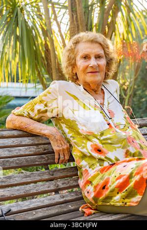 Femme âgée souriante se relaxant sur un banc en bois au milieu d'un jardin tropical vibrant, trempant dans l'atmosphère paisible et la lumière chaude du soleil, incarnant le Banque D'Images