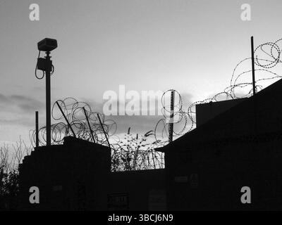 Image monochrome de bobines enchevêtrées de fil de fer barbelé sur un mur en silhouette et caméra de sécurité contre un ciel nuageux du soir Banque D'Images