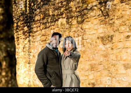 Un couple joyeux se tient devant un mur de pierre rustique, pointant vers quelque chose au loin. Ils sont habillés chaudement, profitant d'une journée ensoleillée ou Banque D'Images