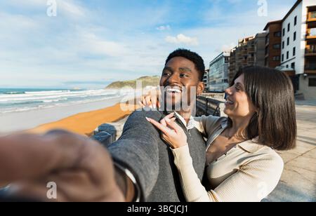 Heureux couple prend un selfie ensemble, souriant et profitant de l'atmosphère de la plage. Avec les vagues de l'océan, le rivage sablonneux et un fond urbain, ils capturent Banque D'Images
