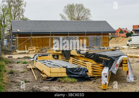 Progression de la construction de maison en bois d'un étage avec du bois empilé en premier plan et structure de construction d'échafaudage dans un quartier résidentiel en développement Banque D'Images