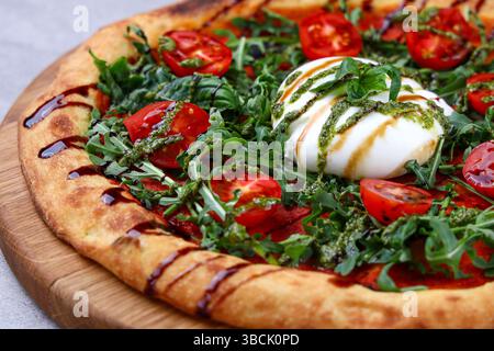Pizza artisanale garnie de burrata crémeuse, roquette fraîche, tomates cerises, pesto et filet balsamique. Banque D'Images