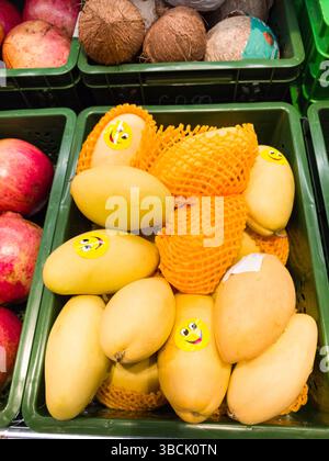Les mangues jaune vif avec des autocollants souriants joyeux sont empilées ensemble dans un panier de marché vert, entouré d'autres fruits frais comme les pommes et co Banque D'Images