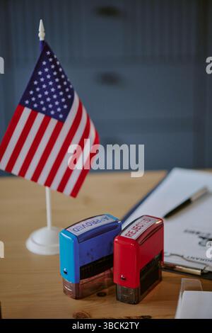 Drapeau américain debout sur un bureau en bois à côté de timbres auto-encreurs rouges et bleus, avec presse-papiers contenant des documents en arrière-plan, créant une configuration de bureau Banque D'Images