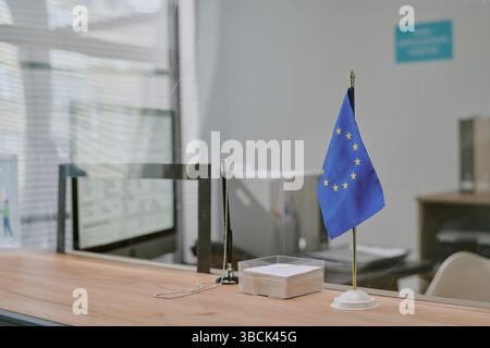 Drapeau de l'UE debout sur le bureau dans un cadre de bureau moderne avec reflet visible dans la fenêtre et arrière-plan flou. Espace de travail vide avec boîte à papier blanche Banque D'Images