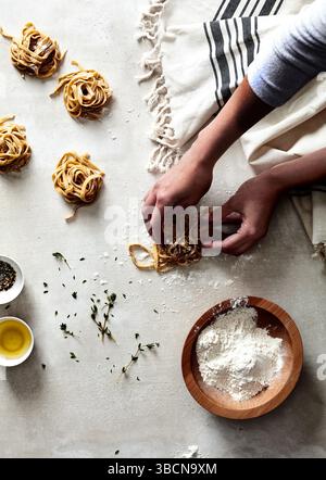 Mains façonnant des nids de pâtes fraîches sur une surface farinée avec des herbes Banque D'Images