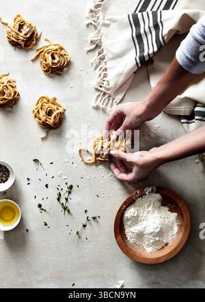 Mains façonnant des nids de pâtes fraîches sur une surface farinée avec des herbes Banque D'Images