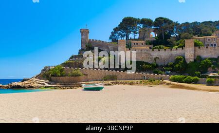 Vue ensoleillée sur les murs du château de Tossa de Mar donnant sur une plage de sable avec un bateau, sous un ciel bleu clair en Catalogne, Espagne. Banque D'Images