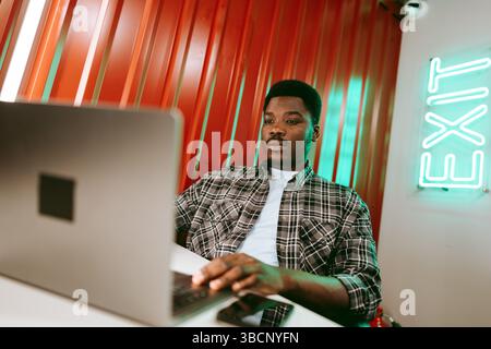 Jeune homme travaillant sur ordinateur portable dans un espace de travail moderne avec des murs rouges vibrants et un panneau de sortie lumineux le soir Banque D'Images