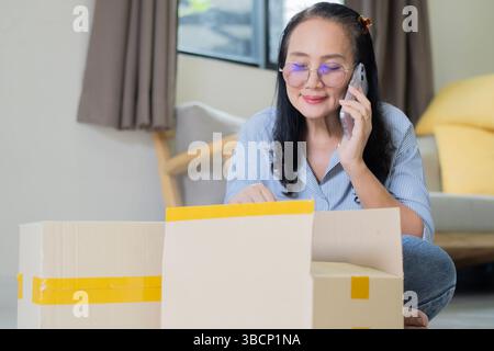 Femme cliente mature positive déballer la boîte de papier à la maison, obtenir l'achat à partir d'un magasin Internet, tenir et utiliser un smartphone. Banque D'Images