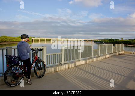 Une personne portant un casque se tient debout avec son vélo sur un pont surplombant une rivière calme pendant l'heure dorée. Banque D'Images