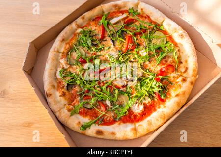 Pizza aux légumes frais avec roquette dans une boîte en carton sur une table en bois, vue de dessus, lumière du soleil. Banque D'Images