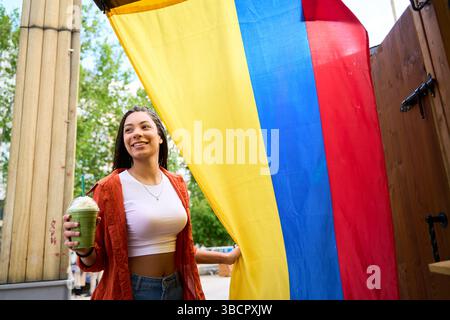 Heureuse jeune femme avec des tresses tenant un grand drapeau colombien, profitant d'un latte matcha à l'extérieur Banque D'Images