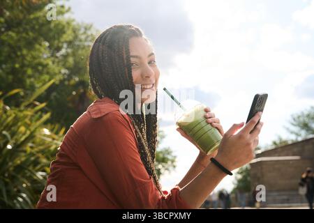 Jeune femme souriante avec des tresses profitant d'un smoothie vert rafraîchissant tout en utilisant son smartphone dans un parc ensoleillé Banque D'Images