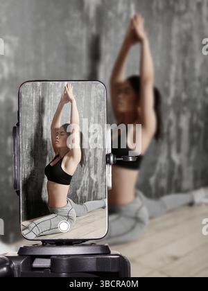 Influenceur de fitness faisant de l'entraînement à domicile et des étirements sur tapis de yoga tout en diffusant en direct sur smartphone. Jeune femme sportive filmant le fitness en ligne Banque D'Images