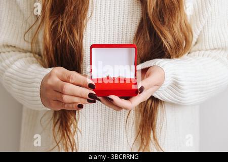 Femme tenant une boîte à anneaux rouge. Boîtier à bijoux ouvert avec bague dorée à l'intérieur. Concept de proposition de mariage. Idée cadeau romantique. Mains élégantes avec manucure sombre Banque D'Images