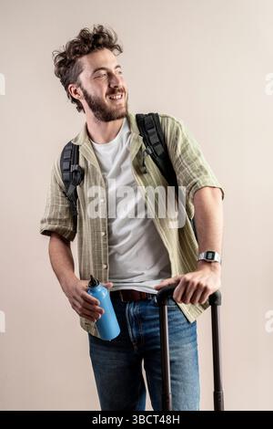 Portrait d'un joyeux homme millénaire tenant une bouteille d'eau réutilisable et valise roulante tout en portant un sac à dos. Studio tourné sur fond neutre Banque D'Images