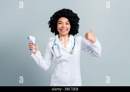 Jeune médecin féminin positif avec les cheveux bouclés tenant un thermomètre et montrant un geste pouce vers le haut sur un fond gris Banque D'Images