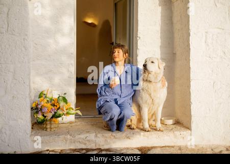 Un moment serein partagé entre une femme en lin bleu et son fidèle chien blanc, profitant d'un lever de soleil paisible à la porte d'une maison traditionnelle trullo dans les Pouilles, en Italie Banque D'Images