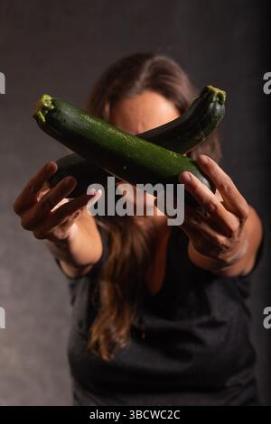 Une femme tient deux courgettes fraîches devant elle. Les courgettes sont vert foncé et disposées en croix. L'accent est mis sur les courgettes, Wit Banque D'Images