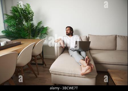 Un homme travaille de façon décontractée de la maison tout en se prélassant sur un canapé moderne dans un intérieur minimaliste et confortable avec des plantes et de la lumière naturelle, symbolisant la détente Banque D'Images