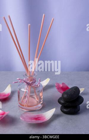 Un diffuseur de roseau est placé sur une surface grise à côté de pierres noires lisses et de pétales de rose dispersés, créant une atmosphère apaisante. Banque D'Images