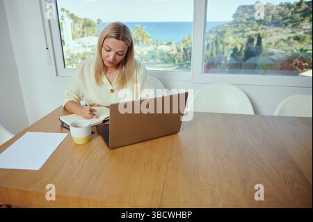 Une femme est assise à une table avec un ordinateur portable et un carnet de notes, travaillant de chez elle dans un espace minimaliste et serein offrant une vue panoramique. Représente le travail à distance, p. Banque D'Images