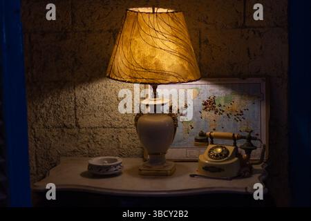 Lampe de chevet diffusant une lueur chaude dans une pièce faiblement éclairée la nuit, avec un téléphone vintage et une carte du monde en arrière-plan Banque D'Images