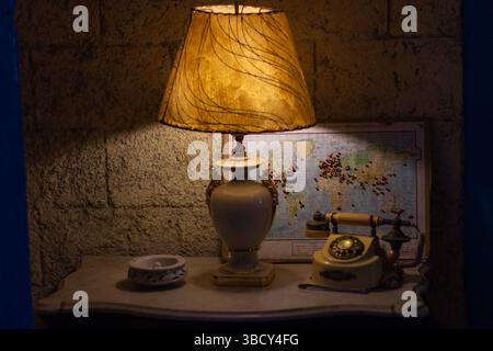 Lampe de chevet diffusant une lueur chaude dans une pièce faiblement éclairée la nuit, avec un téléphone vintage et une carte du monde en arrière-plan Banque D'Images