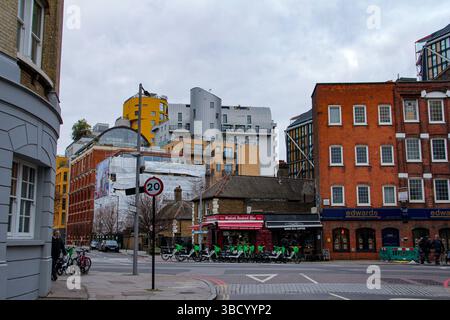 Architecture urbaine variée à Londres mettant en vedette des bâtiments en briques rouges, des appartements modernes, des vélos locatifs et un panneau de limitation de vitesse à 20 mph sous un ciel gris. Banque D'Images